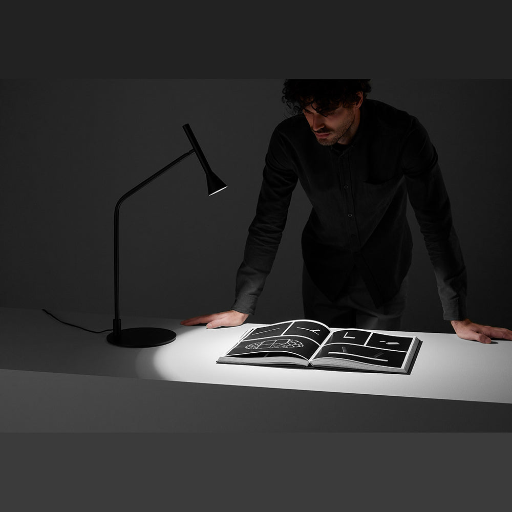 Lyb Table Lamp lit, casting light on an open book while a man leans over a desk in a dark room