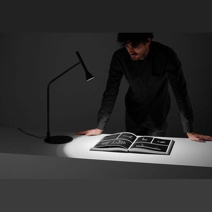 Lyb Table Lamp lit, casting light on an open book while a man leans over a desk in a dark room