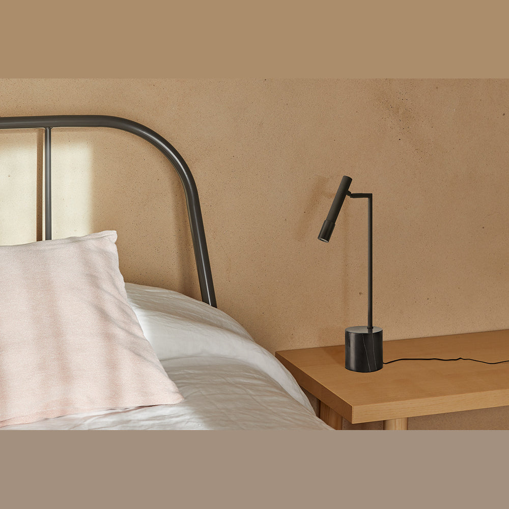Ycro Table Lamp in black on a wooden bedside table beside a bed with a curved metal headboard