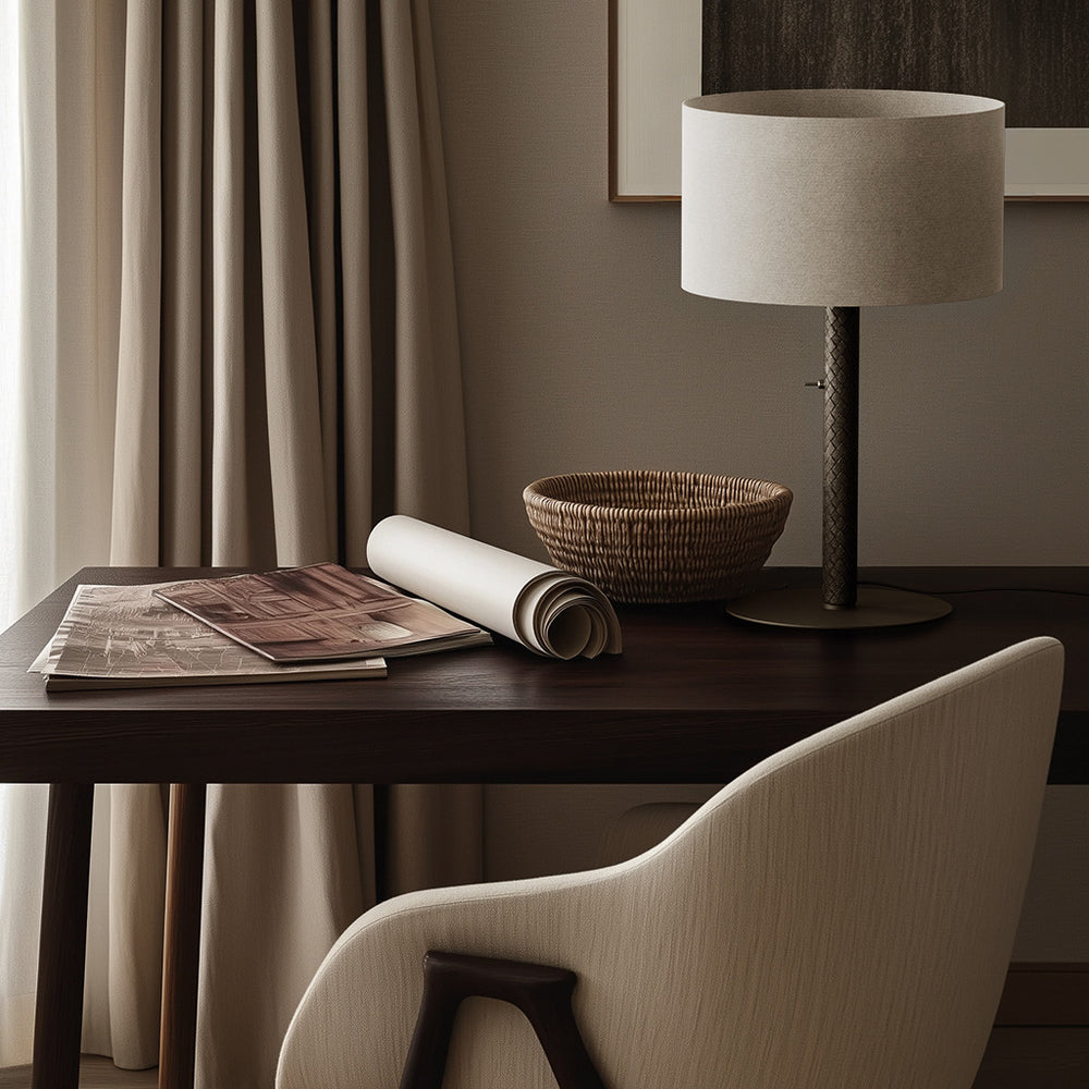Coco Table Lamp with dark stem on a wooden desk beside a woven basket, rolled papers and an upholstered chair